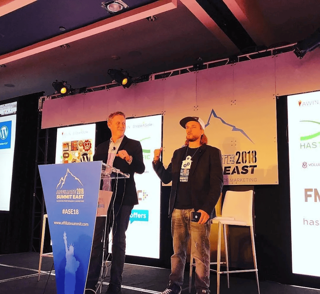David Vogelpohl at the podium and Brian Messenlehner, in a SchoolPresser shirt and cap, on the Affiliate Summit East 2018 keynote stage