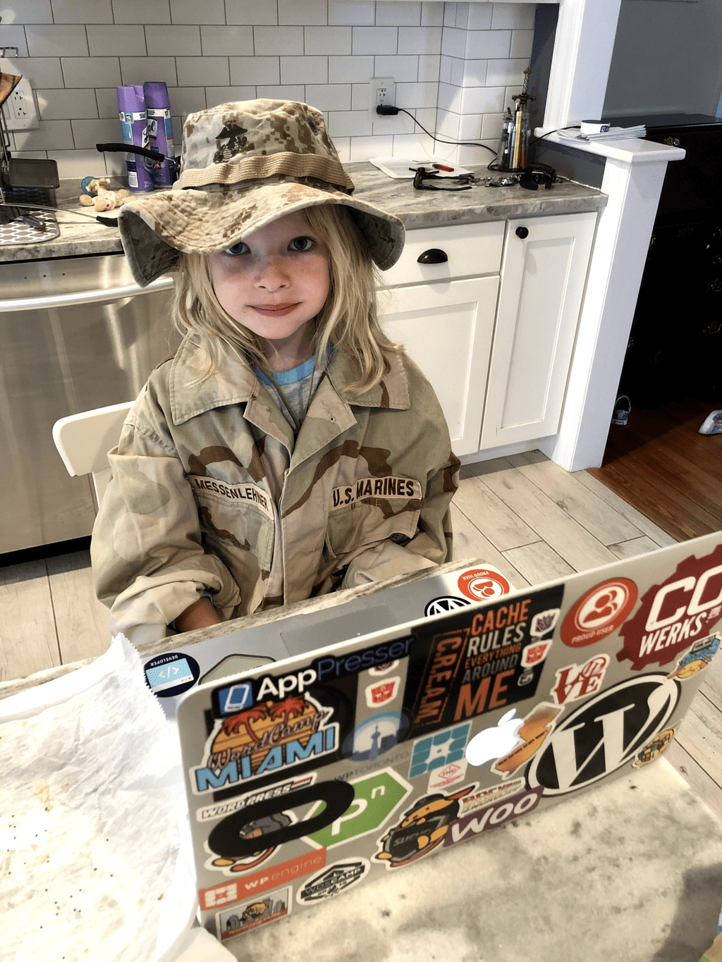 My young daughter wearing my desert camouflage U.S. Marines blouse and boonie hat at a laptop covered in WordPress and AppPresser stickers