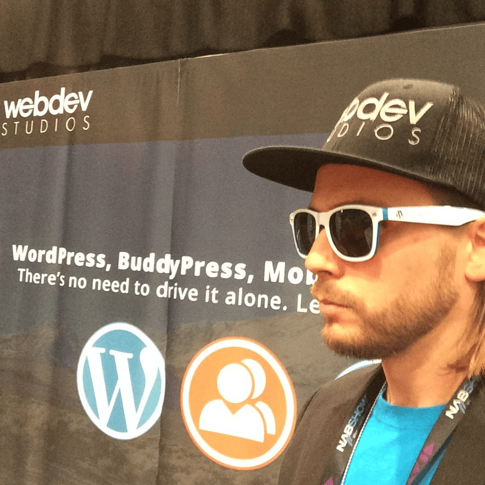 Brian Messenlehner in a WebDevStudios hat, sunglasses and a NAB Show lanyard in front of a WebDevStudios WordPress and BuddyPress banner