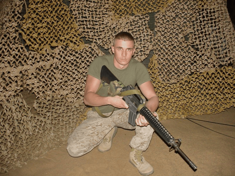 Brian Messenlehner kneeling with his rifle in front of desert camouflage netting, wearing a green T-shirt and desert camouflage trousers