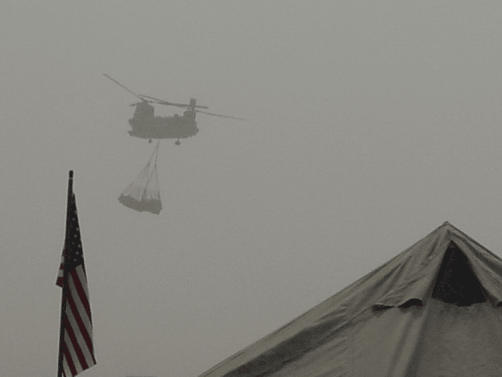 A tandem rotor helicopter carrying a sling load through hazy desert sky over camp, with an American flag and a tent roof in the foreground