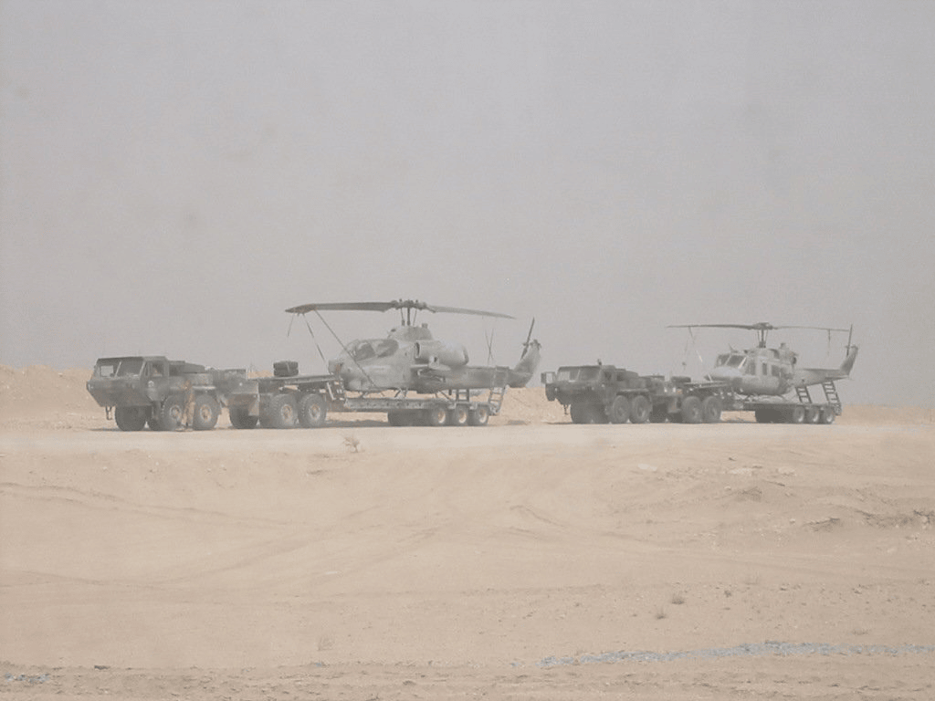 Two Marine Corps helicopters loaded on flatbed trailers behind military trucks in a dusty desert
