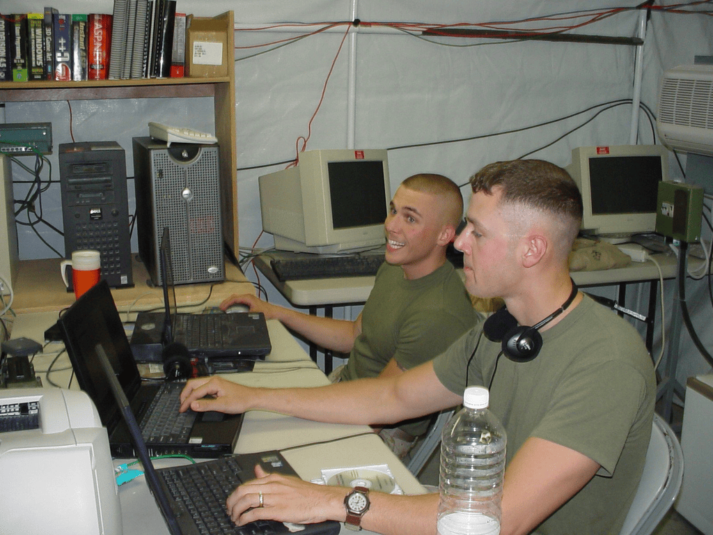 Two Marines in green T-shirts working on laptops in a computer tent, with server towers and monitors around them