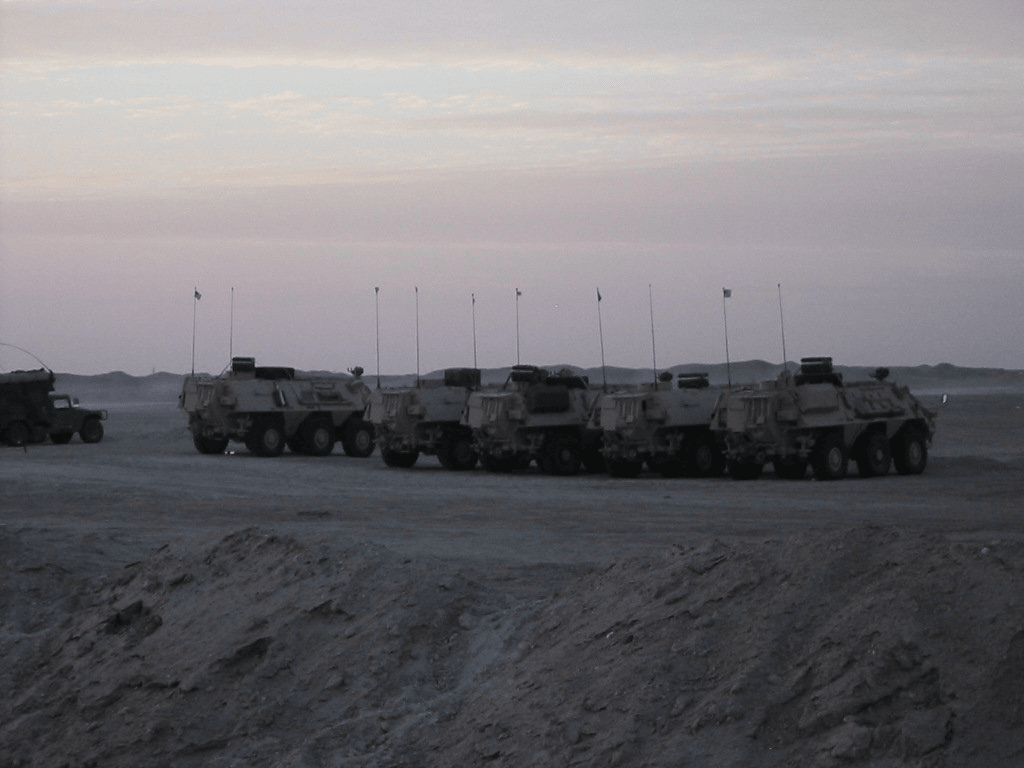 A row of six-wheeled armored vehicles with tall radio antennas parked in the desert at dusk