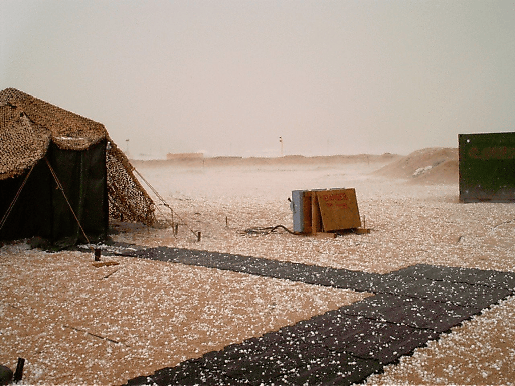 A tent under camouflage netting and a path of ground mats in a desert camp, with the sand covered in hail, during Operation Iraqi Freedom