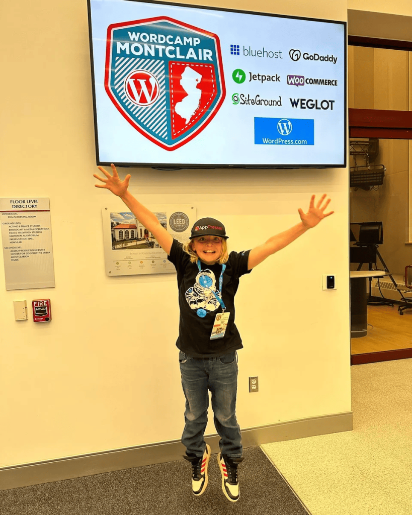 A young attendee celebrating in front of the WordCamp Montclair sponsor banner